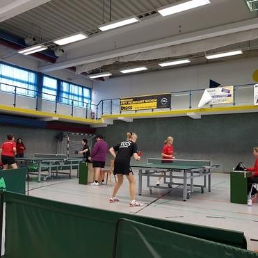 Zwei Tischtennisspielerinnen im Trainingsspiel, eine Spielerin beobachtet das Geschehen am Tisch. Sporthalle im Hintergrund.