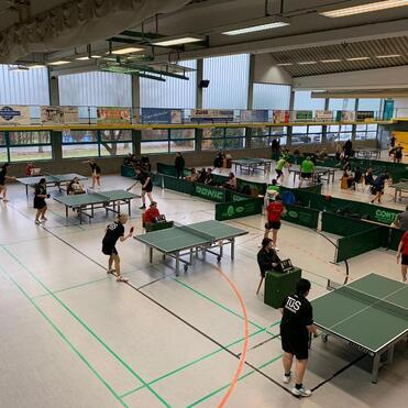 Mehrere Tischtennisspielerinnen und -spieler trainieren in einer großen Sporthalle an verschiedenen Tischen.