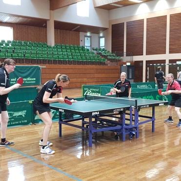 Vier Frauen spielen Tischtennis an einem Tisch in einer Sporthalle mit Holzfußboden und grünen Tribünen im Hintergrund.