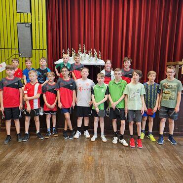 Gruppe von Tischtennisspielern in Sportkleidung steht vor Pokalen auf einer Bühne mit rotem Vorhang im Hintergrund.