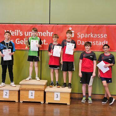 Sechs junge Tischtennisspieler stehen auf Treppchen, halten Urkunden und Pokale vor einem Banner der Sparkasse Darmstadt.