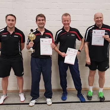 Vier männliche Tischtennis-Spieler stehen nebeneinander, einer hält den Pokal, andere zeigen Urkunden in einer Sporthalle.