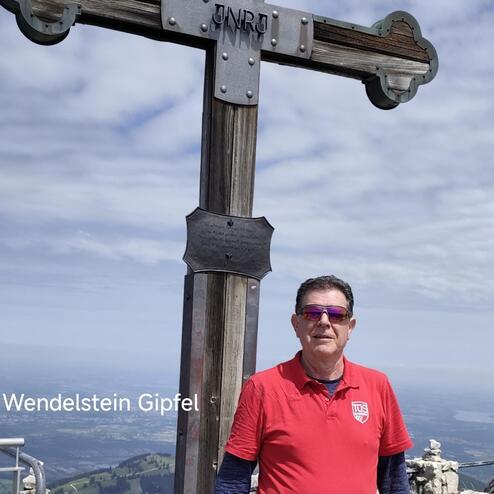 Mann in rotem Shirt steht am Wendelstein-Gipfel vor einem Holzkreuz mit Metallverzierungen und Aussicht auf die Berge.