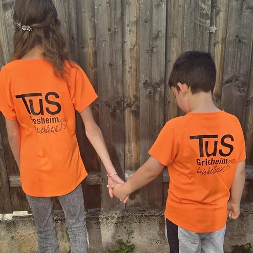 Zwei Kinder, ein Mädchen und ein Junge, stehen Hand in Hand vor einer Holzlattenzaun, beide tragen orangefarbene T-Shirts.