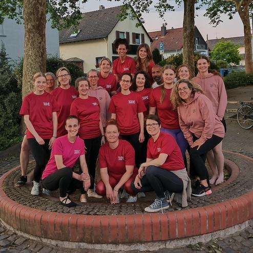 Gruppe von 20 Personen in roten T-Shirts steht auf runder Mauer in einem Park, umgeben von Bäumen und Häusern.
