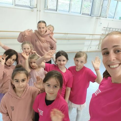 Gruppe von zehn Tänzerinnen in pinken Shirts winkt lächelnd in einem Tanzstudio. Helle Fenster und Ballettstangen im Hintergrund.