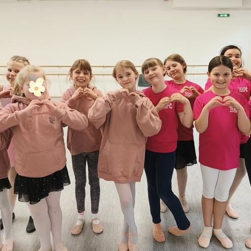 Gruppe von jungen Tänzerinnen in pinken Shirts und Hoodies, die Herzformen mit ihren Händen bilden.