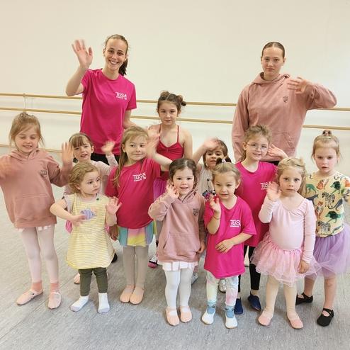 Gruppe von zwölf Kindern und zwei Tanzlehrern in Ballettoutfits, die fröhlich in einem Tanzstudio winken.