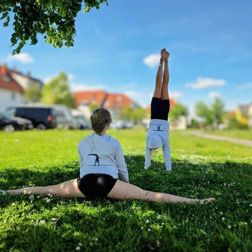 Zwei Mädchen üben Gymnastik im Freien: Eins in Spagat, das andere macht einen Handstand auf einer Wiese.