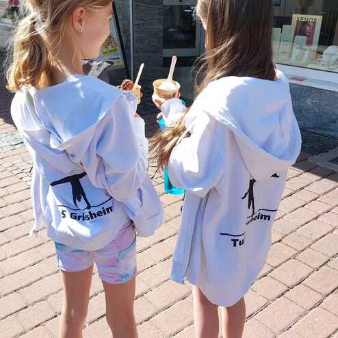 Zwei Mädchen in weißen Kapuzenpullis halten Eisbecher in einer Fußgängerzone auf einem gepflasterten Boden.