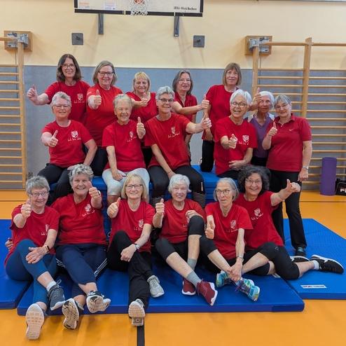 Gruppe älterer Frauen in roten T-Shirts gibt Daumen hoch, posiert in einer Sporthalle auf blauen Turnmatten.