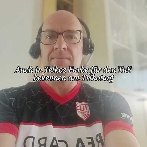 Mann mit Brille und Kopfhörern trägt ein schwarzes Fußballtrikot mit roten Akzenten und ist im Innenraum fotografiert.