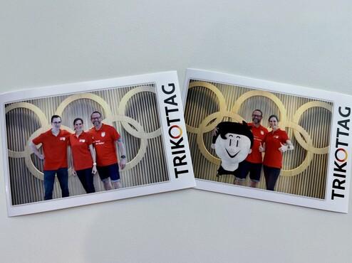 Drei Personen in roten T-Shirts stehen vor einer Wand mit olympischen Ringen, darunter eine Person im Maskottchenkostüm.