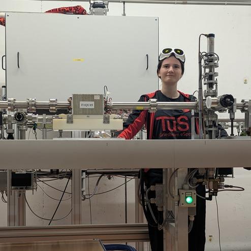 Junge Frau mit Sonnenbrille steht neben einer technischen Apparatur in einem Labor. Kabel und ein Steuerfeld sind sichtbar.