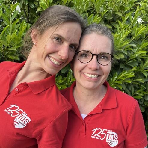 Zwei Frauen in roten Poloshirts mit einem Logo posieren lächelnd vor einer grünen Hecke.