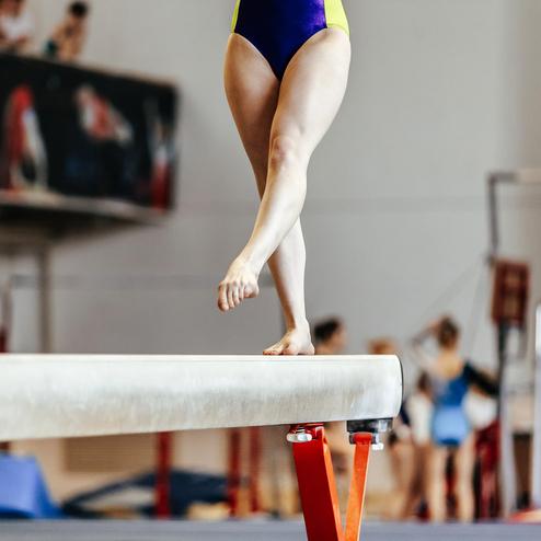 Turnerin balanciert auf einem Schwebebalken in einer Sporthalle, im Hintergrund sind weitere Athletinnen zu sehen.
