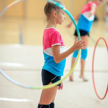 Mädchen in Sportkleidung hält einen hula-hoop-Reifen in einer Turnhalle, im Hintergrund ist ein weiteres Kind sichtbar.