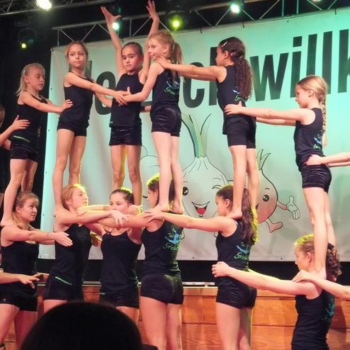 Gruppe von Mädchen in schwarz, die in einer Akrobatik-Darbietung auf einer Bühne stehen, mit buntem Hintergrund.