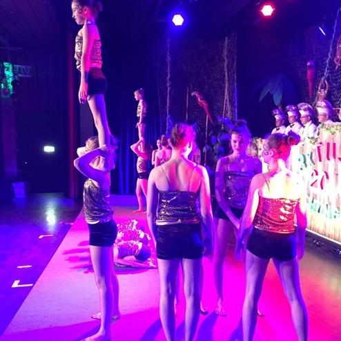 Gruppe von Artisten auf der Bühne, eine Person steht auf den Schultern einer anderen, bunt beleuchtet.