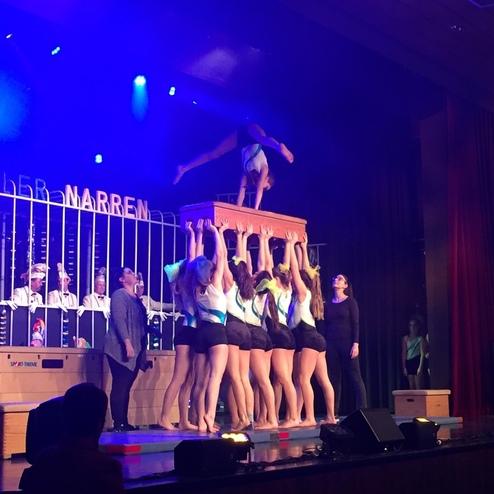 Aufführung mit Tänzerinnen, die eine Pyramide bilden, während eine Person oben auf einer Kiste steht, beleuchtet von bunten Lichtern.