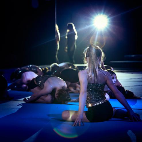 Tänzerin in glitzerndem Top sitzt auf blauer Matte, während andere Tänzerinnen im Hintergrund auf dem Boden liegen.