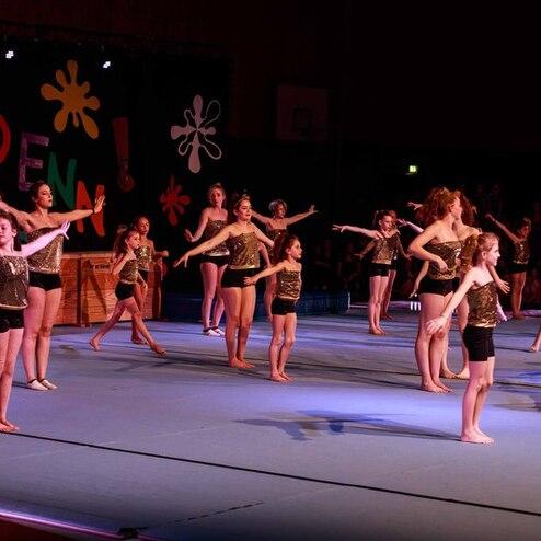 Gruppenvorstellung von Tänzerinnen in goldenen Oberteilen und Shorts auf einer Gymnastikfläche bei einer Aufführung.