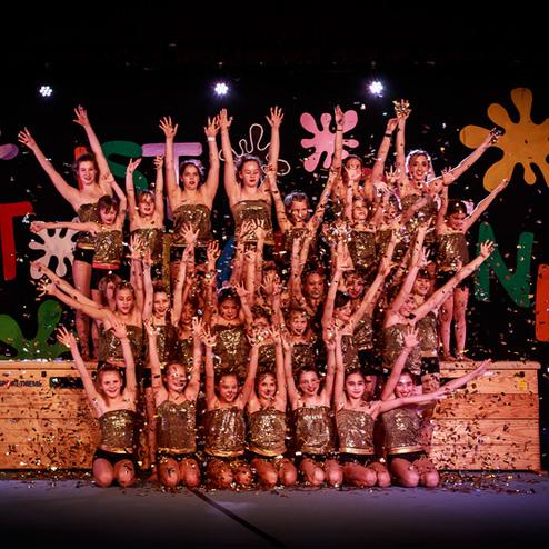 Gruppe von Tänzerinnen in goldenen Kostümen, die auf einer Bühne mit bunten Dekorationen posieren und Konfetti werfen.