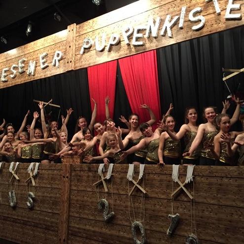 Gruppe von Tänzern in goldenen Kostümen posiert vor einem hölzernen Puppentheater mit roten Vorhängen.