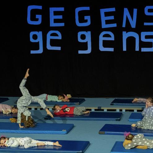 Kinder in Schlafanzügen liegen auf blauen Matten, während eines von ihnen einen Handstand inmitten einer Aufführung macht.