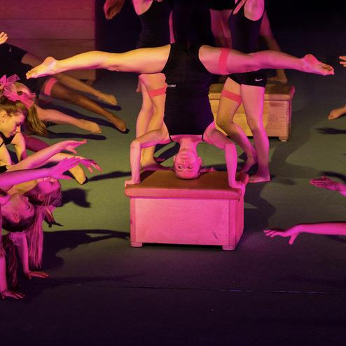 Gruppe von Tänzerinnen in Schwarz führt synchronisierte Bewegungen in pinkem Licht auf einer Bühne aus.