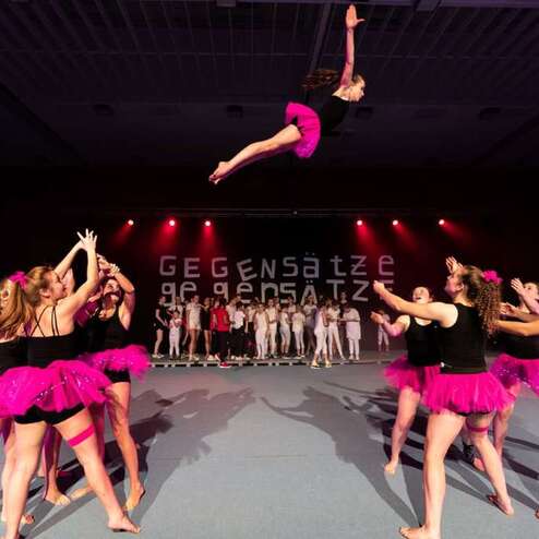 Mädchen in einem pinken Tutu wird von Tänzerinnen auf einer Bühne hochgeworfen, während Zuschauer im Hintergrund applaudieren.