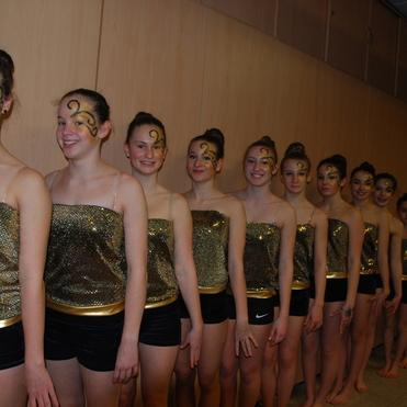 Reihe von Tänzerinnen in glitzernden Outfits mit goldener Gesichtsfarbe stehen in einer Halle.