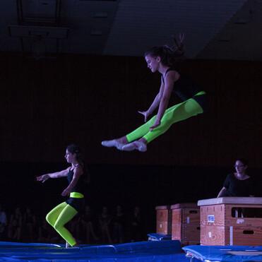 Zwei Turnerinnen in neonfarbenen Leggings springen von Holzbarren auf eine blaue Landematte während einer Trainingssession.