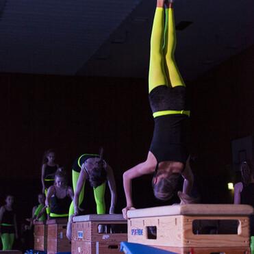 Gymnastinnen führen Übungen an Holzgeräten aus, einige stehen kopfüber, in neonfarbenen Leggings und Oberteilen.