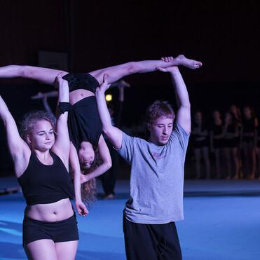 Zwei Artisten heben ein Kind in der Mitte einer Aufführung auf einer Turnfläche, umgeben von weiteren Tänzern.