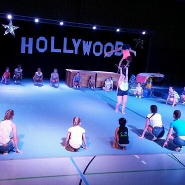 Auf einer Sportfläche führen Akrobaten einen Partnerwurf vor, während Zuschauer um sie herum sitzen. Hintergrund mit "HOLLYWOOD"-Buchstaben.