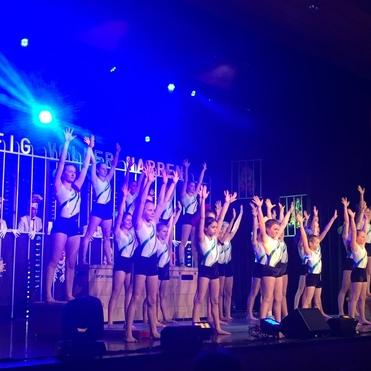 Tanzaufführung mit mehreren Kindern in Sportkleidung, die ihre Arme erhoben auf einer Bühne stehen.