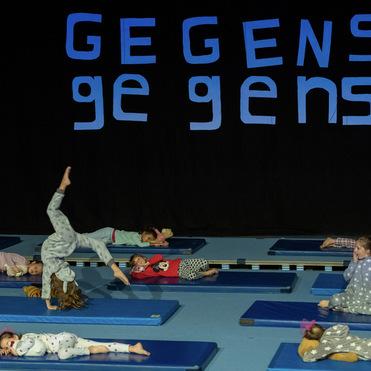 Kinder in Schlafanzügen liegen auf blauen Matten, während eines von ihnen einen Handstand inmitten einer Aufführung macht.