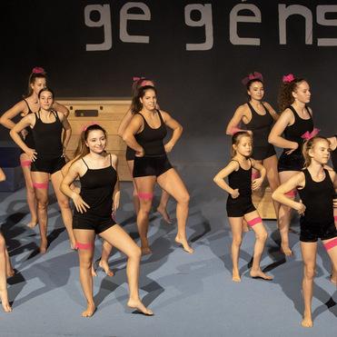 Gruppe junger Tänzerinnen in schwarzen Outfits mit pinken Akzenten, die auf einer Bühne synchron tanzen.