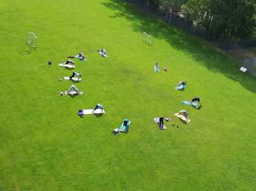 Vogelperspektive auf eine Wiese mit mehreren Personen, die auf Matten liegen und sich entspannen oder trainieren.