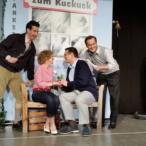 Szenenbild eines Theaters mit vier Schauspielern, die in einem Gespräch auf einer Holzbank sitzen, im Hintergrund eine Zapfsäule.