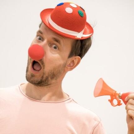 Mann mit roten Clownsnase und Hut hält eine kleine Tröte, überrascht schauend vor einfarbigen Hintergrund.