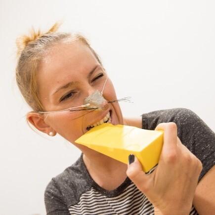 Frau mit einer Mausmaske beißt in ein Stück gelben Käse in einer hellen Umgebung.