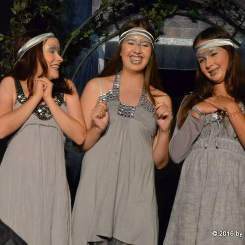 Drei Tänzerinnen in grauen Kleider mit Stirnbändern lächeln vor einer mystischen Kulisse auf der Bühne.