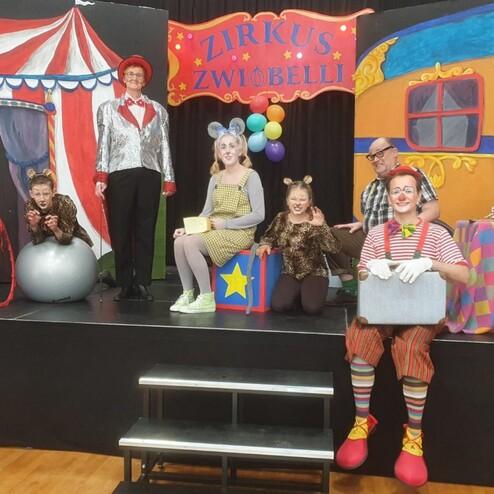 Sechs Personen in Kostümen stehen auf einer Bühne vor einem Zirkushintergrund mit bunten Dekorationen.
