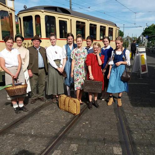 Gruppenausstellung von Menschen in historischen Kleidern mit Körben vor einer alten Straßenbahn an Gleisen.