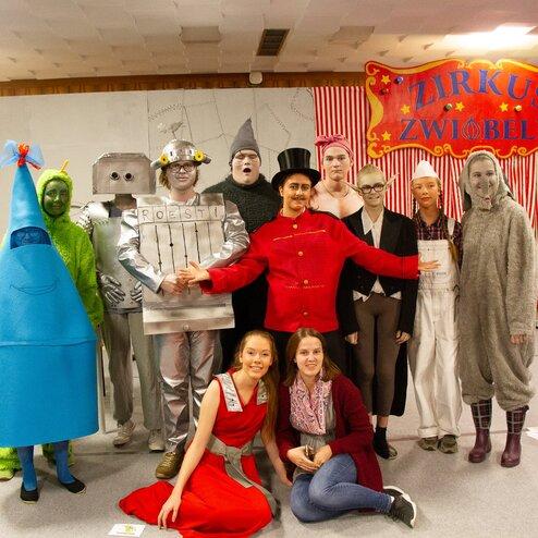 Gruppenszene im Zelt mit verkleideten Darstellern, darunter Zirkusdirektor und vielseitige Figuren in bunten Kostümen.