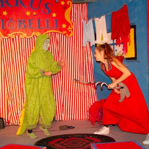 Zwei Schauspieler auf einer Zirkusbühne: einer in grünem Kostüm, der andere in rotem Kleid mit einem Haken.