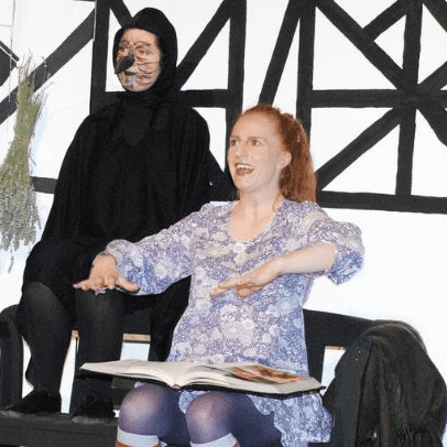 Zwei Schauspieler auf einer Bühne: Eine Frau mit Buch und eine maskierte Figur im Hintergrund vor einer verzweigten Wand.