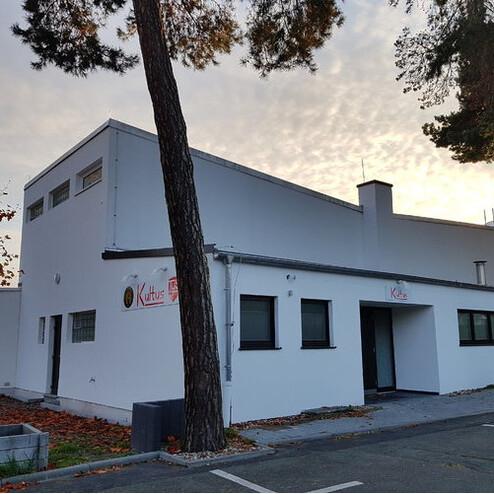 Modernes, weißes Gebäude mit großen Fenstern und einem Eingangsschild, umgeben von Bäumen und herbstlichem Laub.